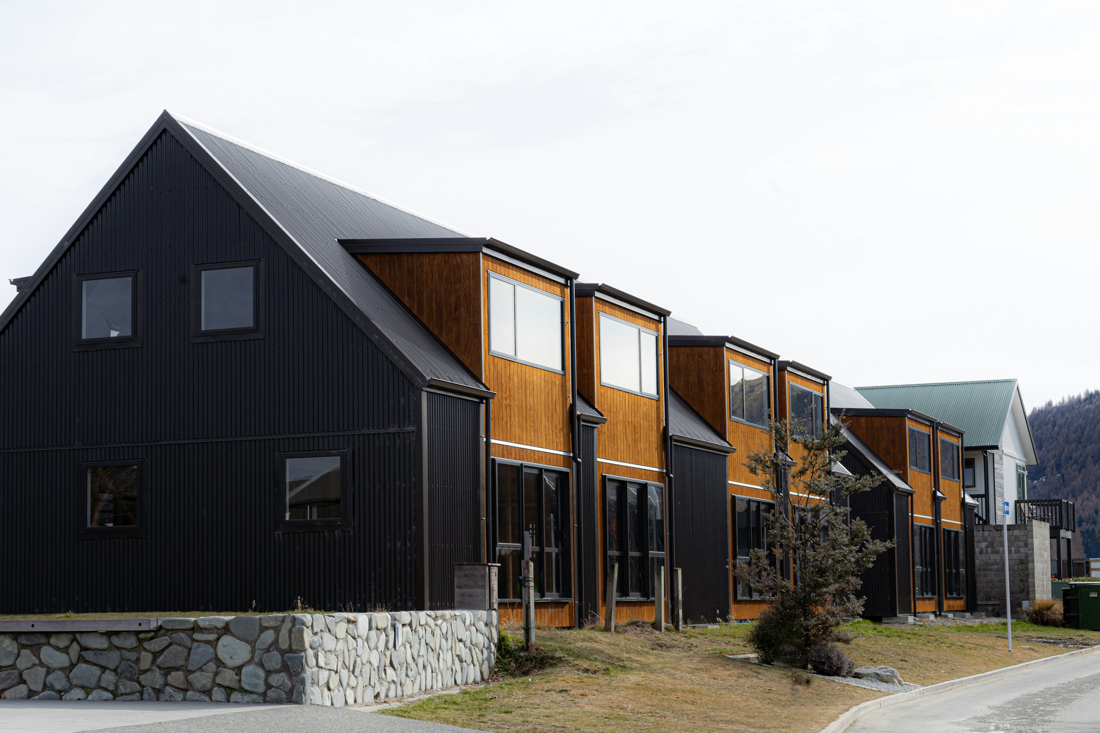 A row of townhomes with dark ribbed metal cladding and warm cedar upper storeys, on a quiet street below bare hills.