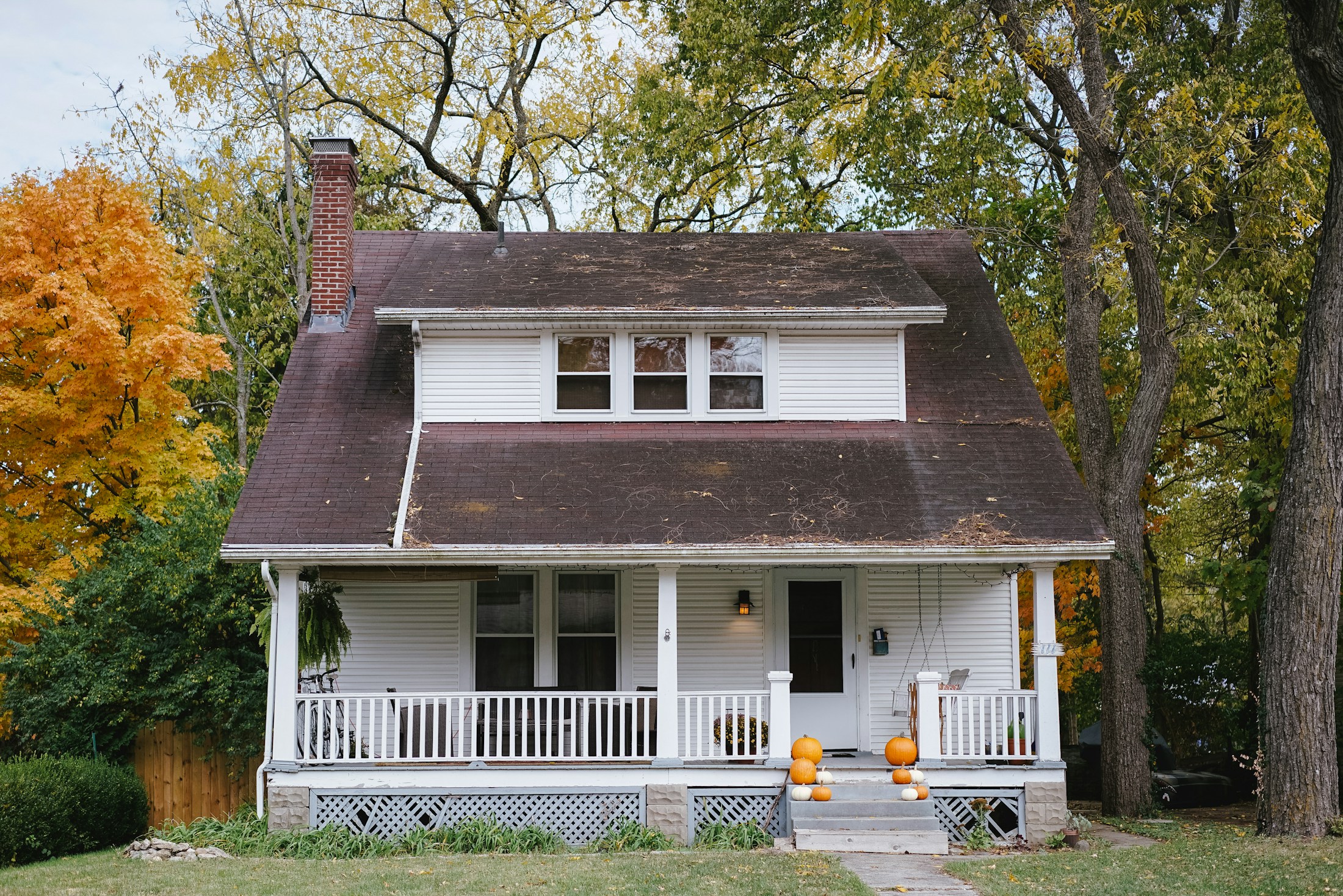A white clapboard house with a deep covered porch, a brick chimney and autumn trees behind it.